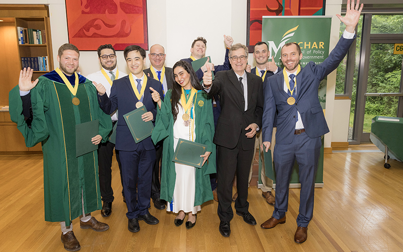 Nine people stand on a wooden floor and wave to the camera, with many of them wearing mortarboards and robes.