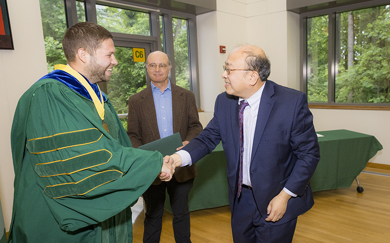 A man in a green robe with a yellow ribbon around his neck shakes hands with a man in a blue suit while a man in a brown jacket watches.