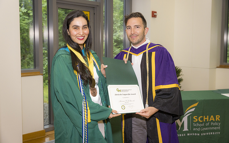 A woman in a green robe with a yellow ribbon around her neck holds a certificate with a man in a purple robe.