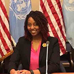 A woman in a black jacket and pink shirt sits in front of 2 American flags