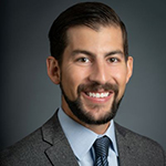 A man in a gray suit, light blue collared shirt, and tie stands and smiles
