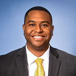 A man in a black suit, white collared shirt, and yellow tie, stands and smiles