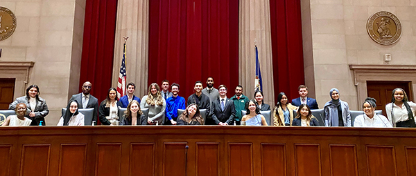 A group of people sit in chairs while others stand behind them in a large, regal room.