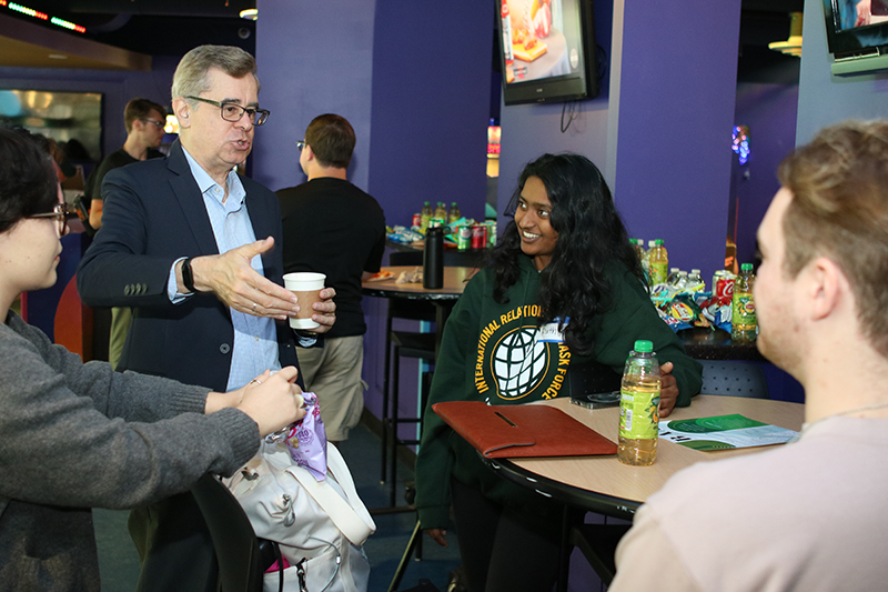 Three students sit listening to a man wearing a blue blazer and blue shirt and holding a cup of coffee.