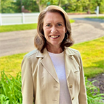 A woman in a cream jacket and white shirt stands outside and smiles