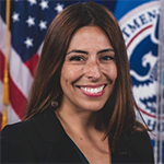 A woman in a black shirt stands in front of an American flag