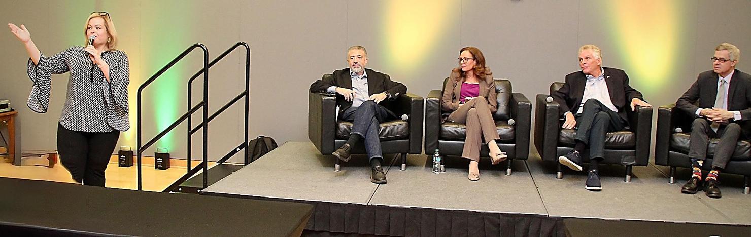Four people sit on a stage while a woman speaks into a microphone.