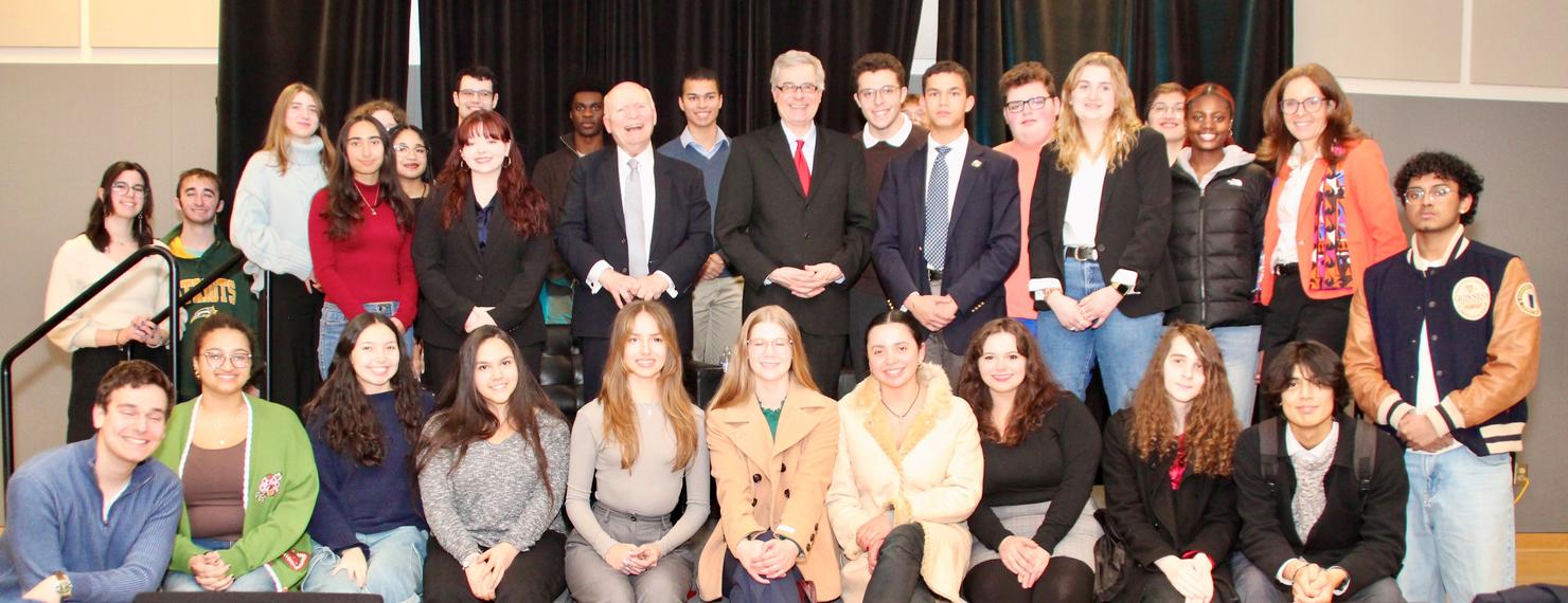 Two men in suit jackets and neckties stand on stage surrounded by smiling college students.