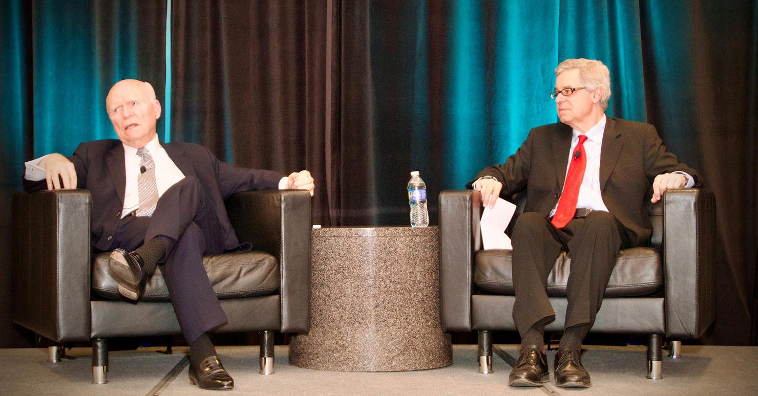 Two men in suit jackets and neckties sit on black chairs in front of blue drapes.