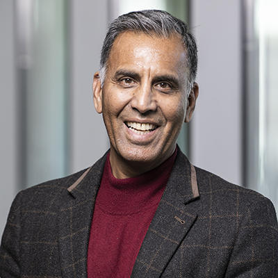 A man wearing a jacket and red collared shirt stands inside while smiling