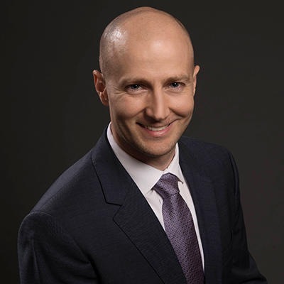 A man in a black suit, white collared shirt, and silver tie stands smiling in front of a black background