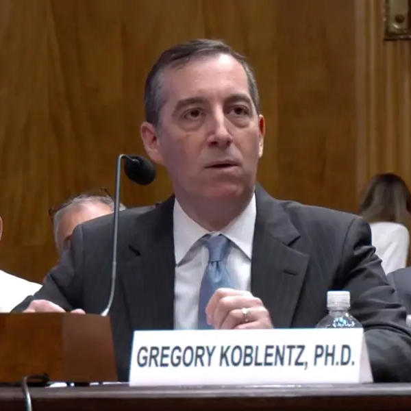 A man in a gray suit and blue tie sits at a desk with a microphone.
