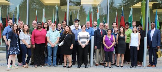 A group of 25 adults who are smiling in front of international flags.