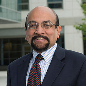 A man in a black suit, white striped shirt, tie, and glasses stands in front of Van Metre Hall and smiles