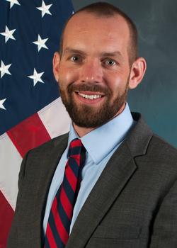 Schar School PhD in Biodefense 2014 PhD graduate Brian Mazanec stands in front of an American flag.