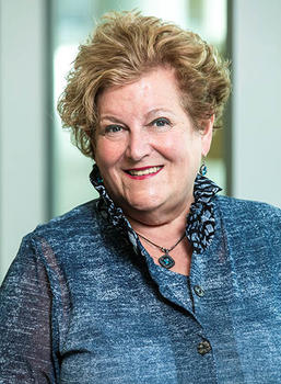 A woman in a blue top with a blue pendant necklace smiles at the camera.