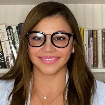 A woman with dark hair and large eye glasses smiles at the camera with a row of books behind her.