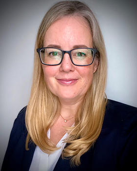 A woman with long blonde hair and eye glasses gazes at the camera.