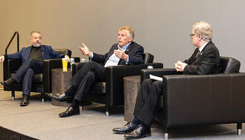 3 men in dark suits sit in leather chairs on a stage.