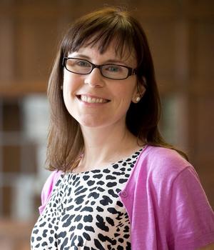 A woman with brown hair and glasses wears a pink sweater and smiles at the camera.