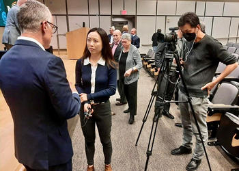 A man in a blue suit stands with his back to the camera as a woman interviews him in front of a video camera.