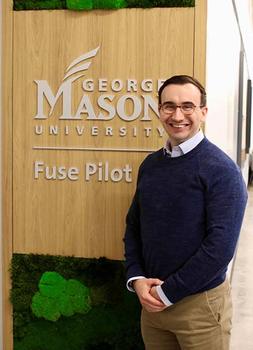 A man in a sweater and eyeglasses stands in front of a sign for a building.