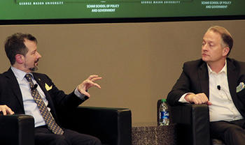 Sean Kirkpatrick and Shane Harris wearing dark jackets sit in black chairs and talk.