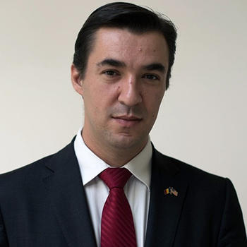 A man with short dark hair in a dark jacket and white shirt gazes at the camera.