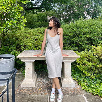 A woman in a white dress leans on a table on a patio surrounded by shrubs.