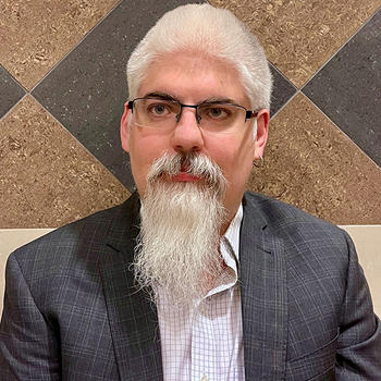 A man with white hair and a pointy white beard and eyeglasses sits in front of a geometric tile wall.