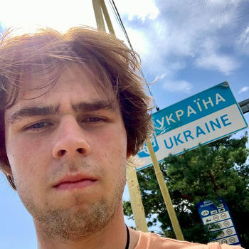 A young man in an orange tee shirt looks at the camera as he stands in front of a Ukrainian road sign.