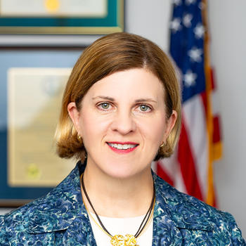 A woman in a blue jacket and with a gold flower necklace stands in front of an American flag.