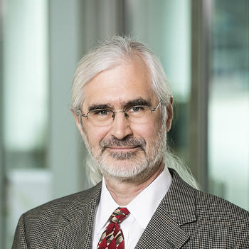 A man with long grayish hair and a beard, wearing glasses and a jacket and tie poses for the camera.
