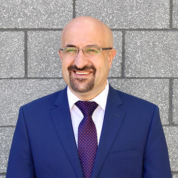 A bald man in eyeglasses, a blue suit, and with a goatee smiles off camera.